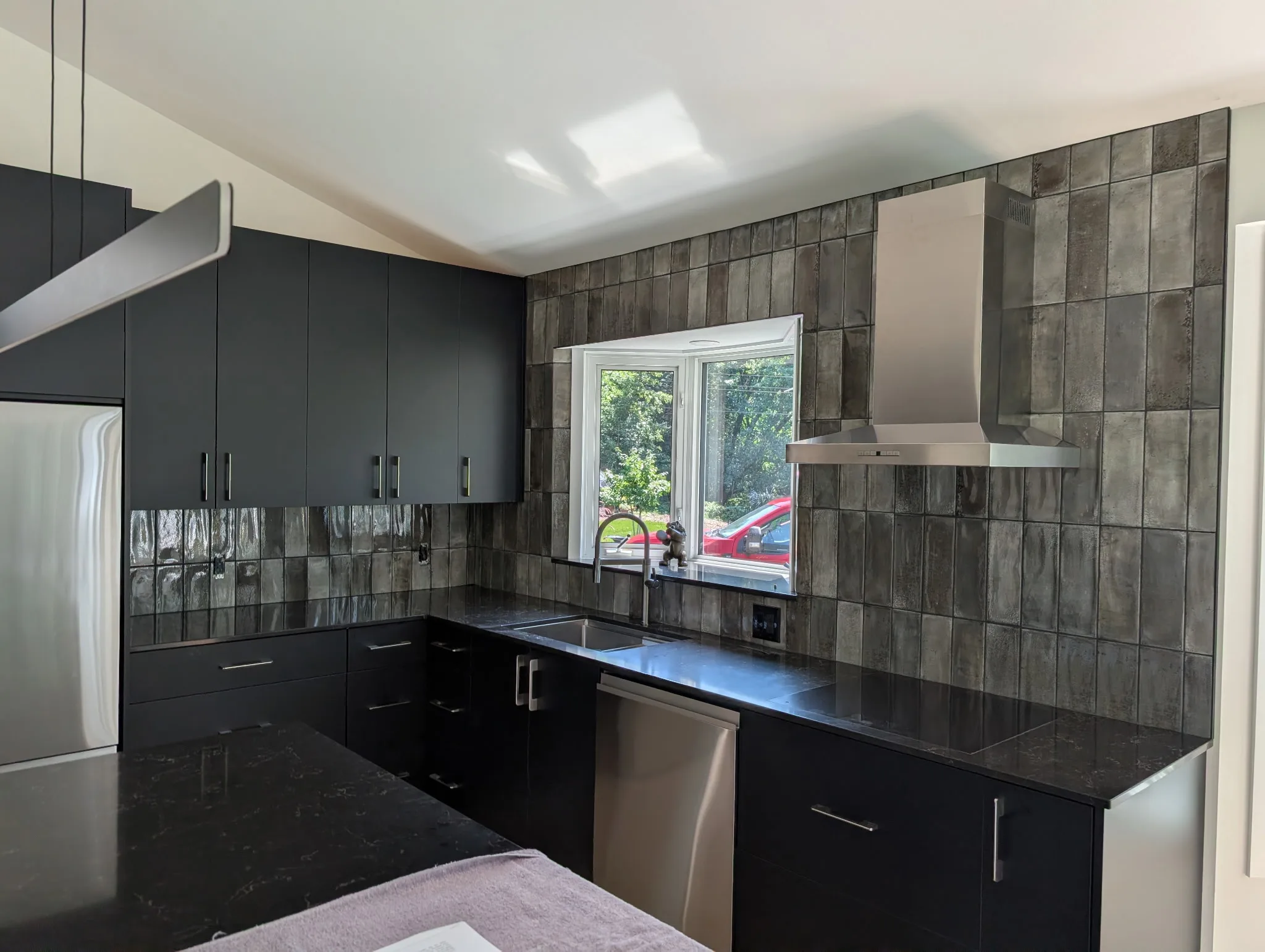 Grey vertical stacked kitchen backsplash tile running floor to ceiling around a window, with black cabinets and quartz countertops, installed by Torino Tile & Stone